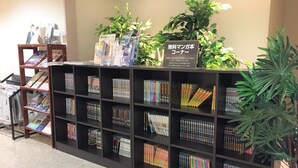 Ein Bücherregal mit verschiedenen Büchern und Zeitschriften, ein Schild, das einen kostenlosen Manga-Bereich kennzeichnet, und einige Blumentöpfe.