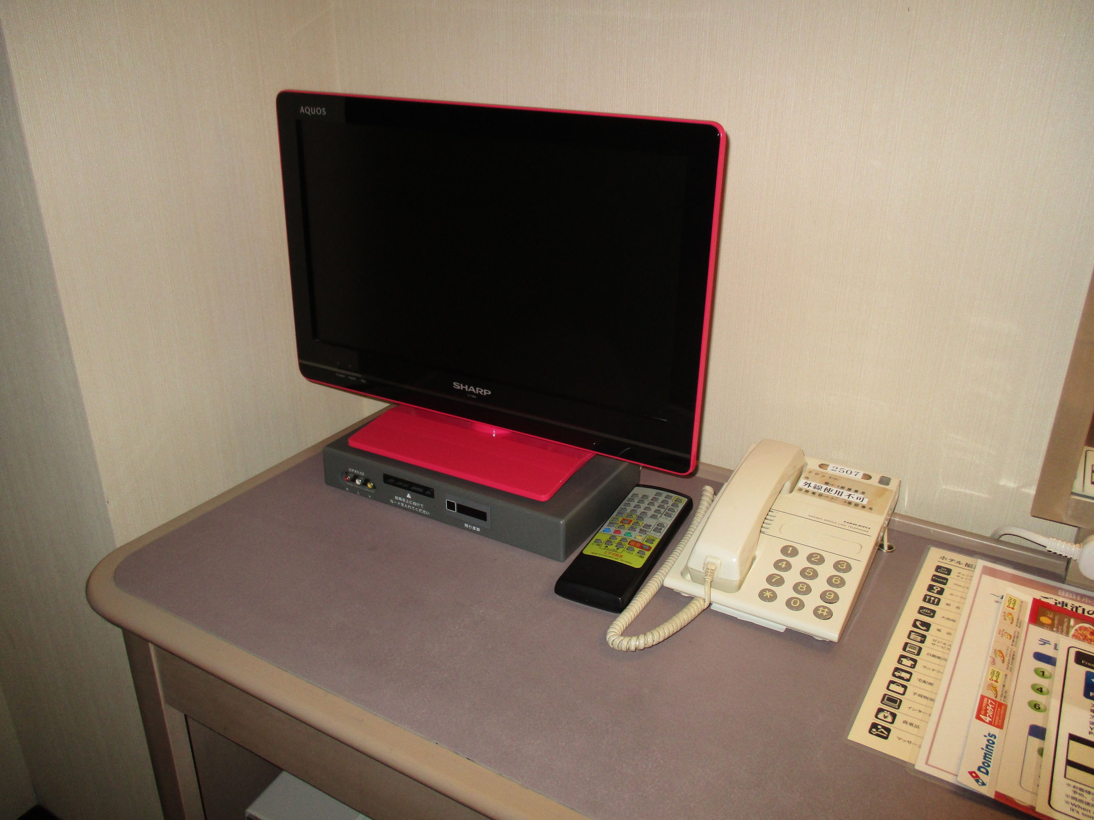 A hotel room with a Sharp monitor, a telephone, and a remote control on a desk.