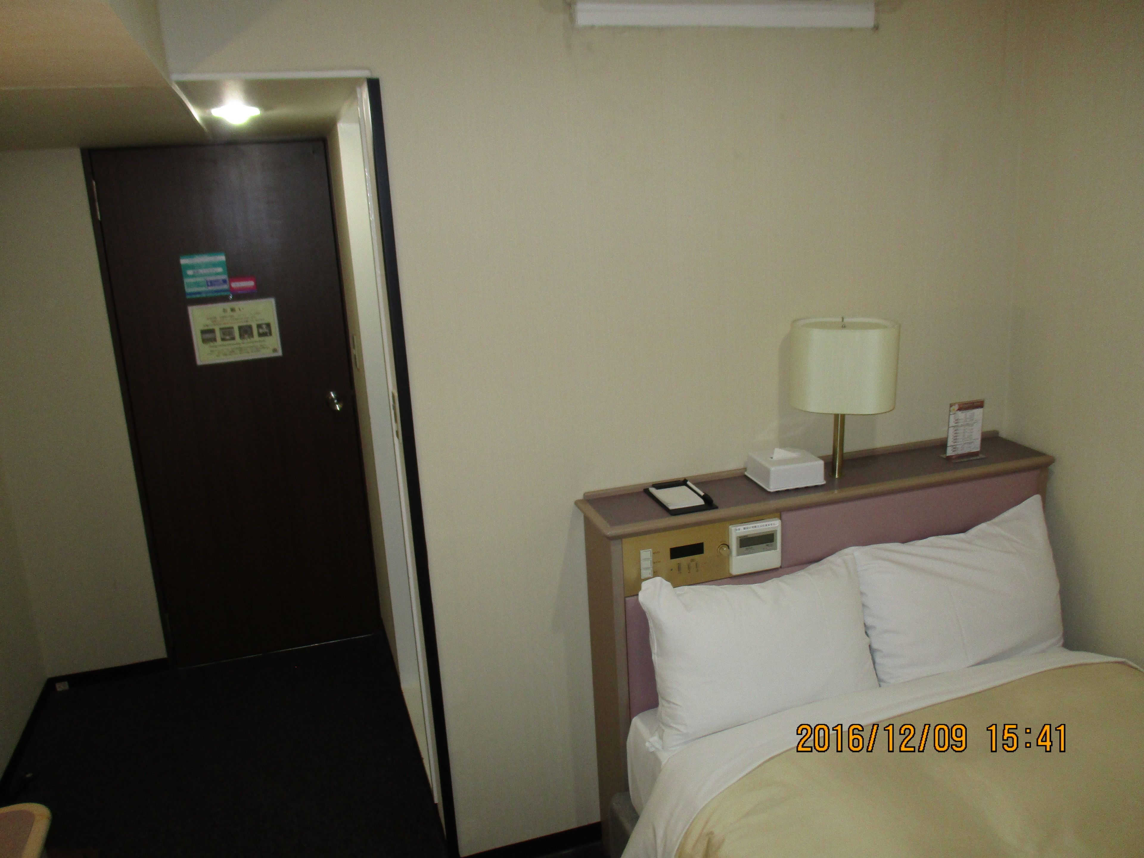 Habitación de hotel con cama, mesita de noche, lámpara y una puerta con un cartel.
