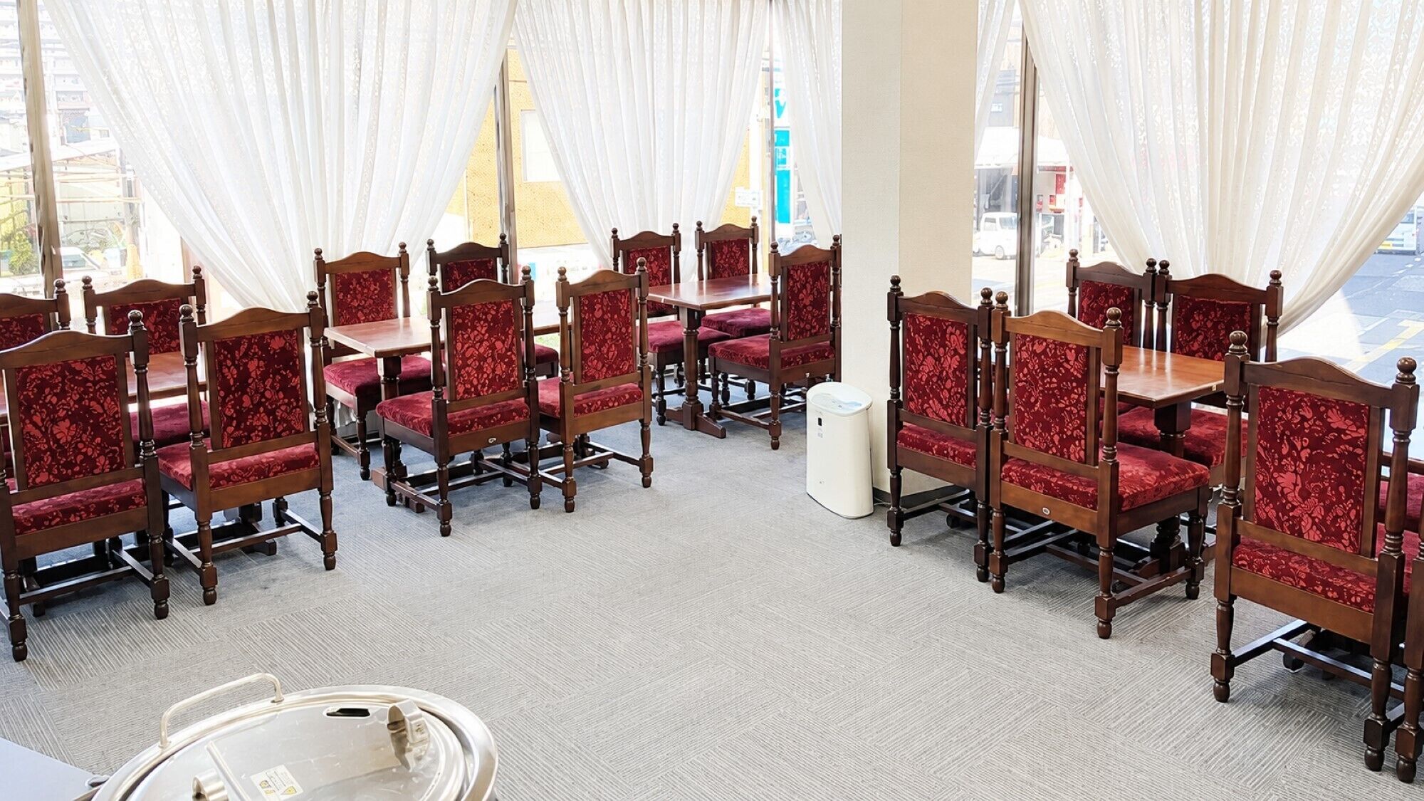 A conference room with wooden chairs and tables, white curtains, and a view of the street outside.