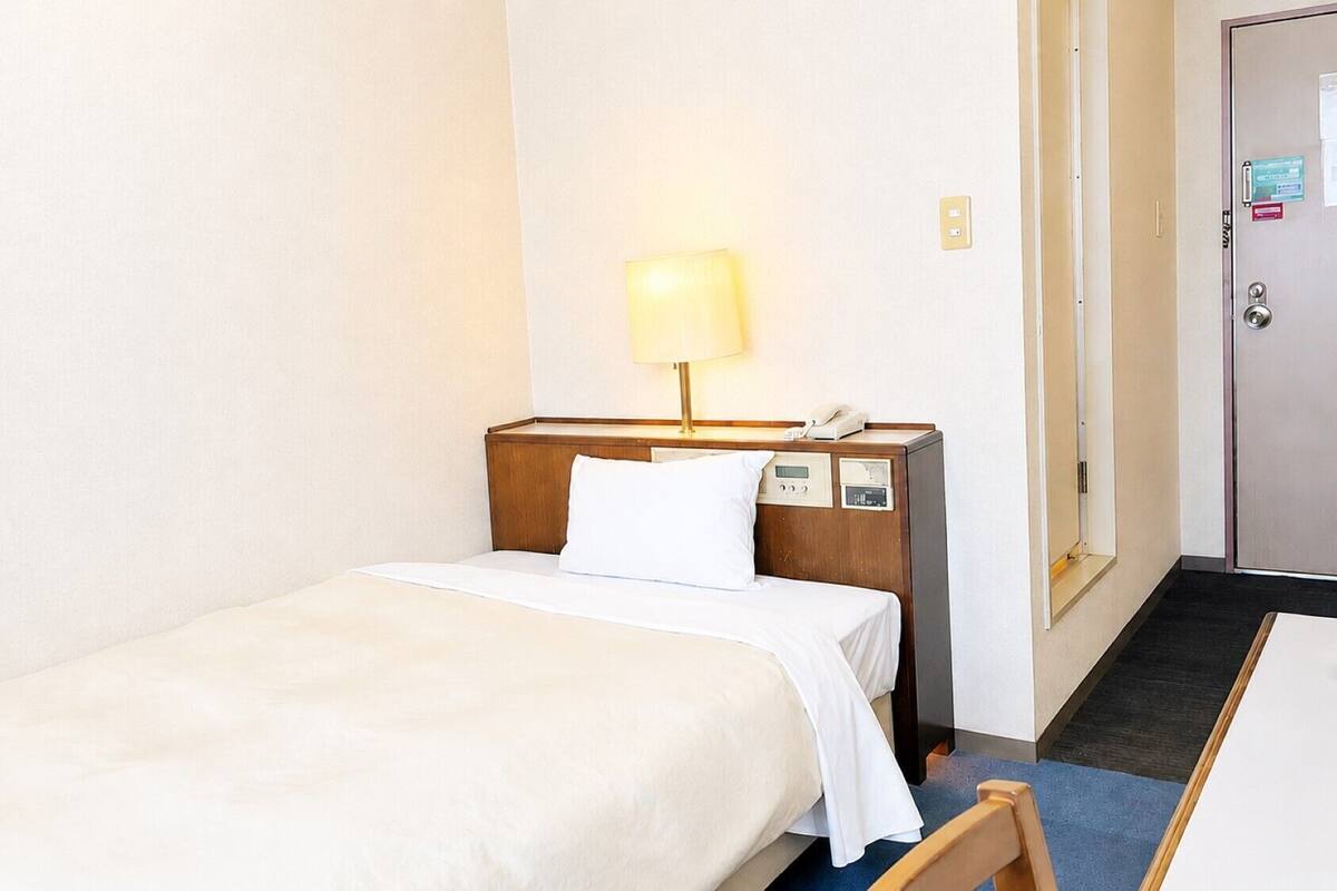 Une petite chambre d’hôtel avec un lit simple, une table de chevet avec une lampe, un téléphone et une porte sur le côté droit.