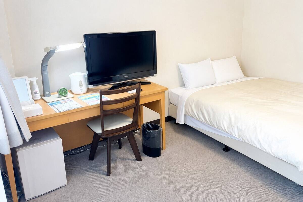 Une chambre d’hôtel avec un lit, un bureau avec un ordinateur, une chaise, une petite poubelle et un radiateur blanc.