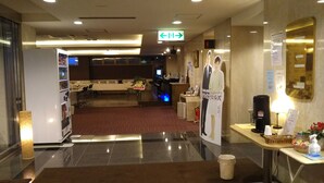 A hotel reception area with a reception desk, a digital screen, a sign indicating an exit, and a counter with amenities. - Hotel Hitachi Hills (Hitachi)