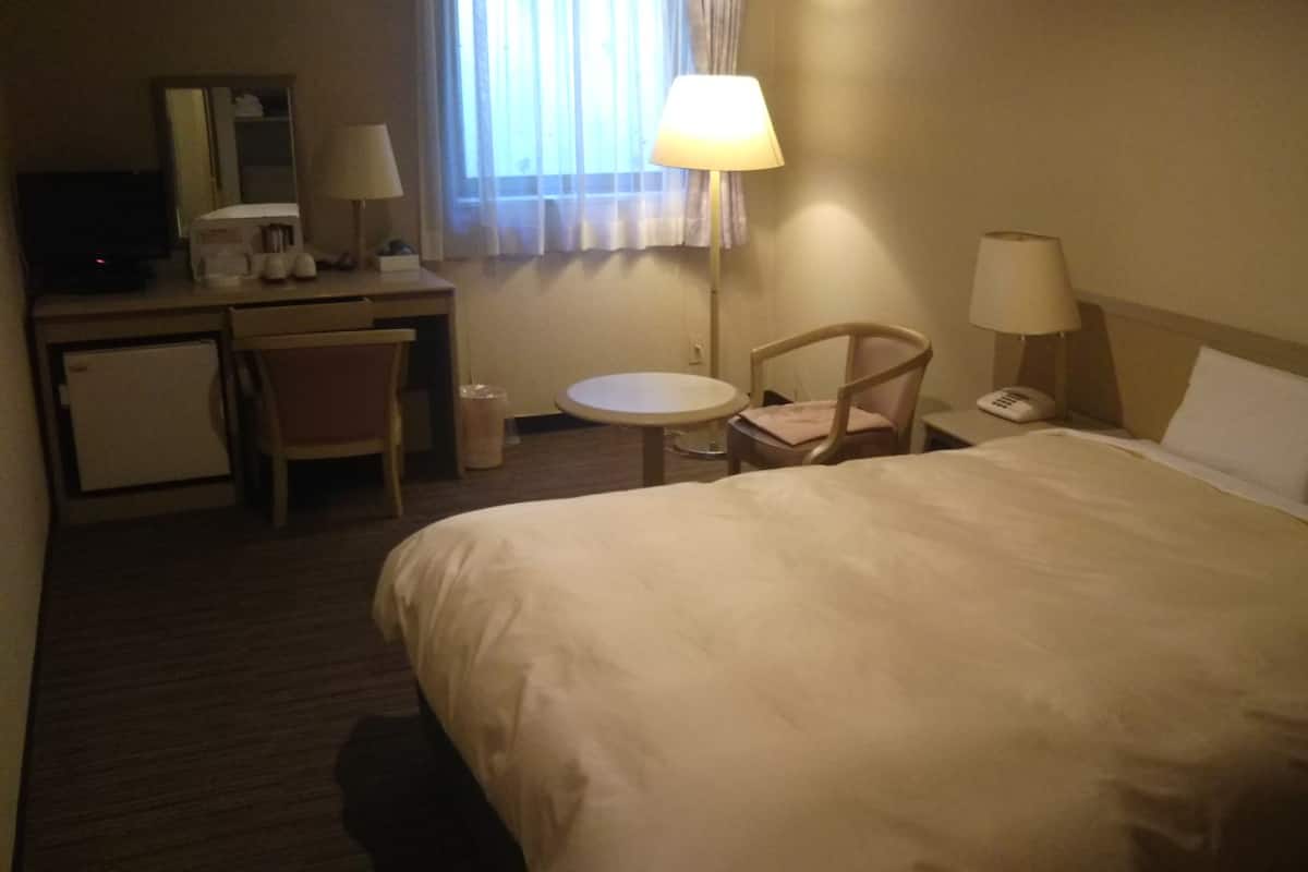 Quarto de hotel com uma cama, uma escrivaninha com abajur, uma cadeira, uma mesinha e um telefone.