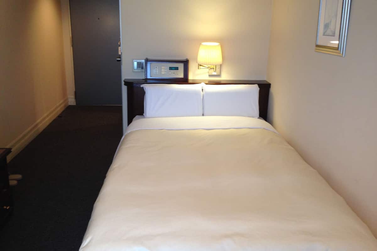 Et hotelværelse med en seng, et natbord med en lampe og en termostat på væggen.