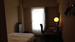 Una habitación de hotel con cama, escritorio, silla, lámpara y ventana con cortinas.