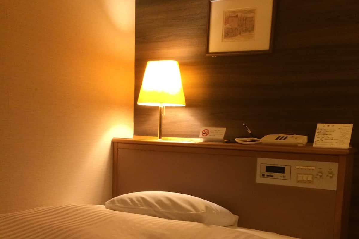A hotel room with a bed, a bedside table with a lamp, a phone, and a control panel.