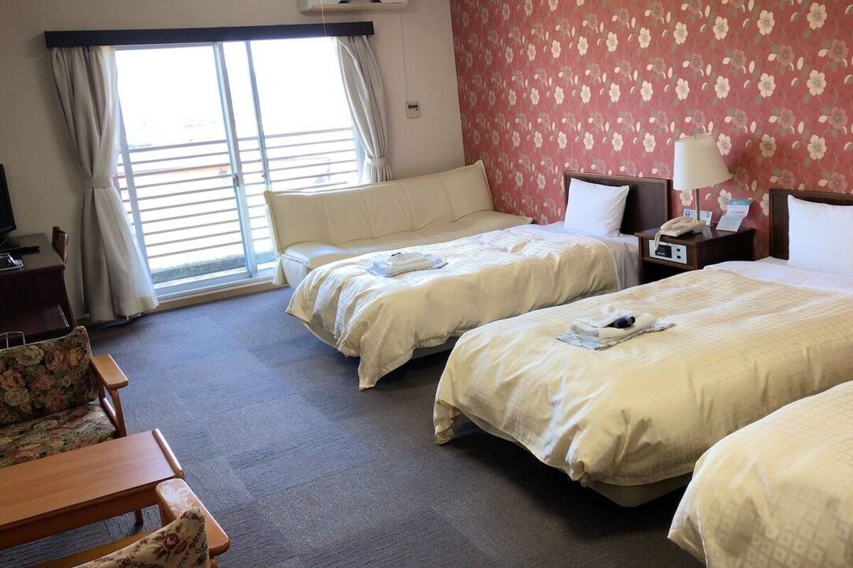 Ein Hotelzimmer mit drei Betten, einer Sitzecke, einem Schreibtisch und einem Fenster mit Vorhängen.