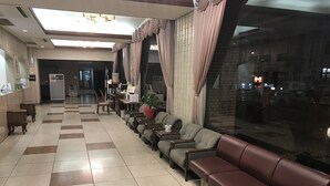 A hotel lobby with a check-in counter, seating area with sofas and armchairs, a reception desk, and a large window offering a view of the street.