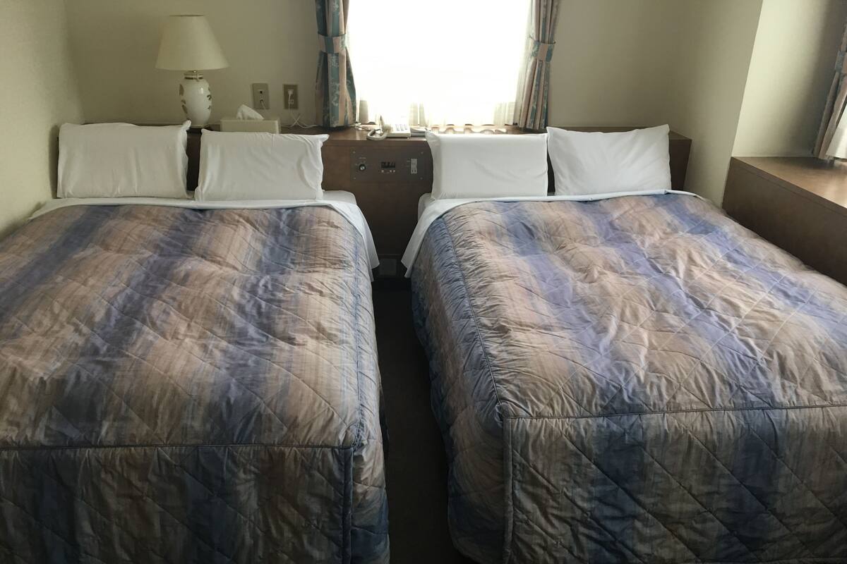 Ein Hotelzimmer mit zwei Betten, jeweils mit gesteppter Bettdecke, einem Nachttisch mit Lampe, einem Telefon und einem Fenster mit Vorhängen.
