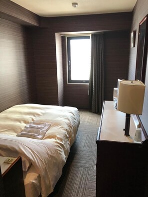 A hotel room with a bed, a desk, a lamp, and a window with curtains. - Hotel Crown Hills Nakamura (Shimanto)