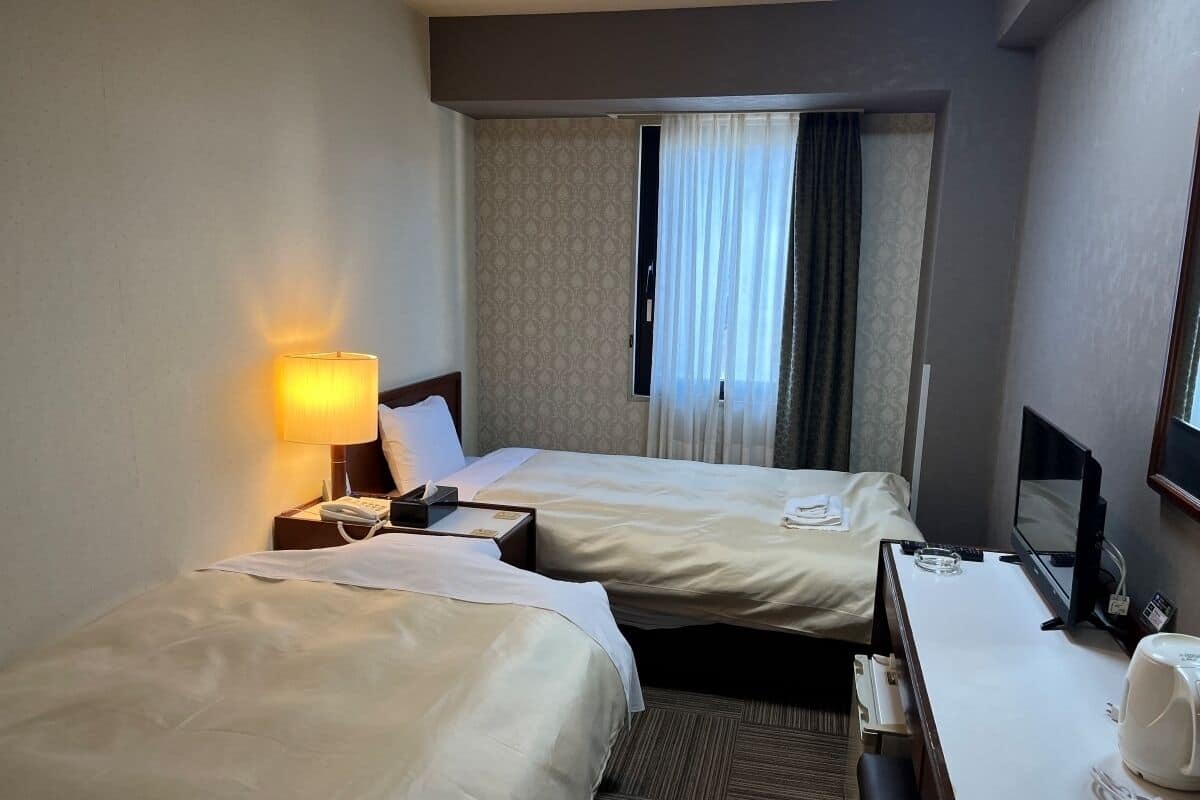 Une chambre d’hôtel avec deux lits, un bureau, une chaise, une lampe, une télévision et une fenêtre avec des rideaux.