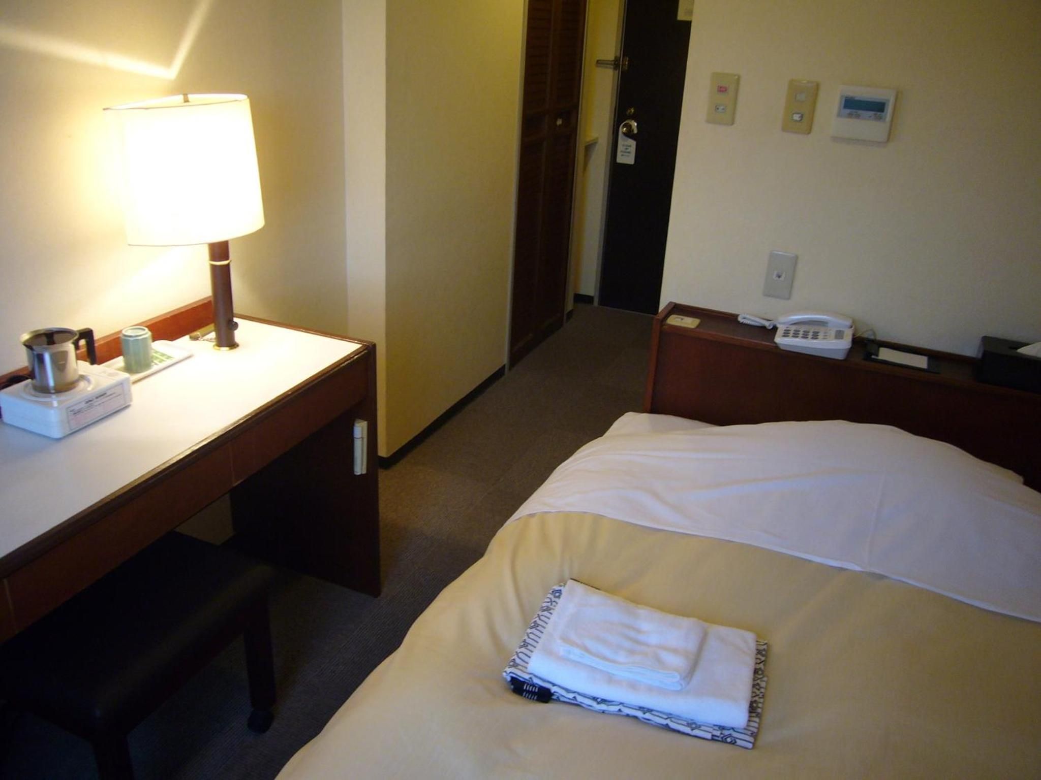 A hotel room with a bed, a desk with a lamp, a telephone, and a wall-mounted thermostat.