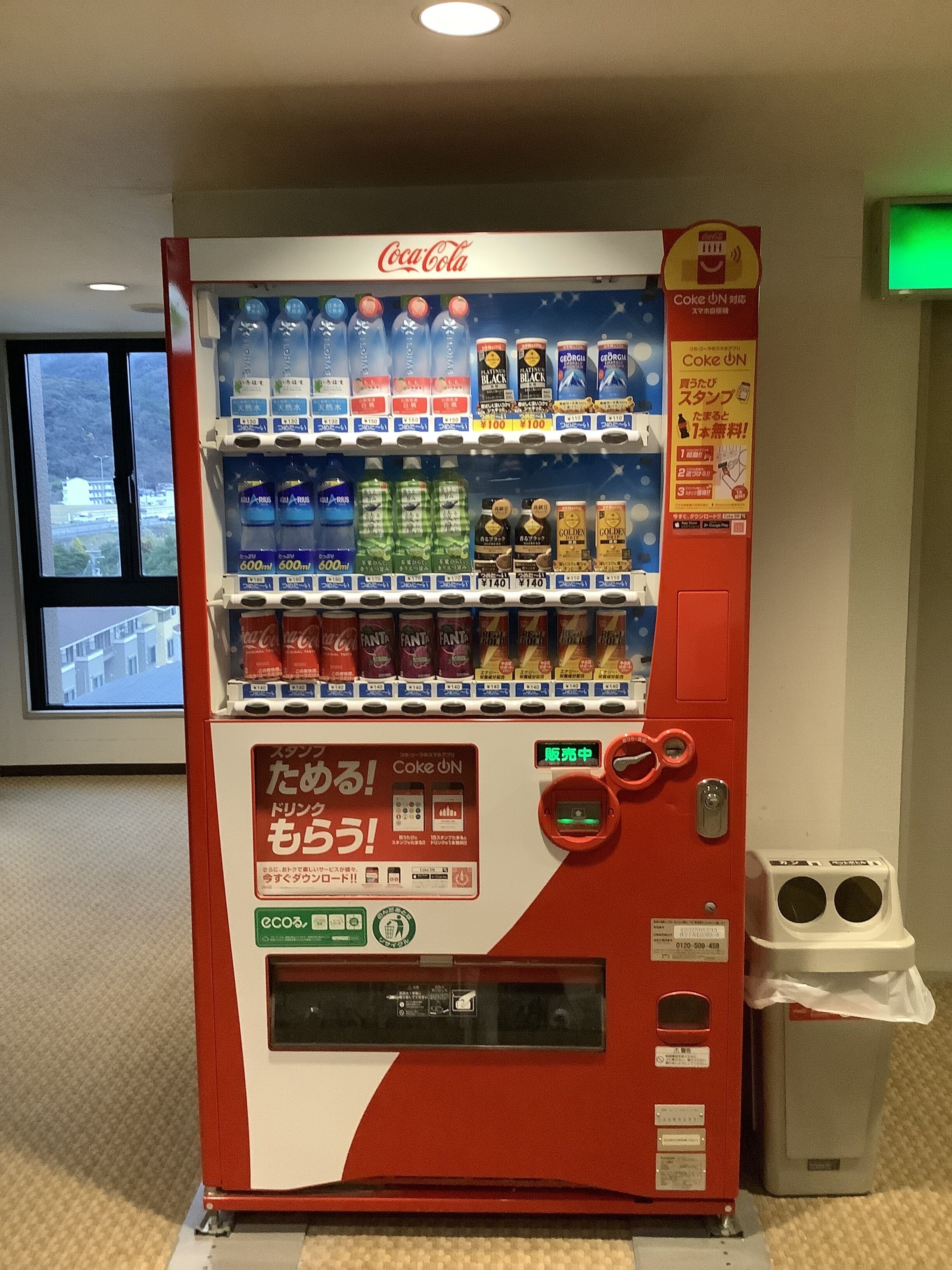 A Coca-Cola vending machine with various drink options and a recycling bin nearby.