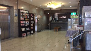 A hotel lobby with a check-in counter, a reception desk, a bookshelf with magazines, and a tiled floor.