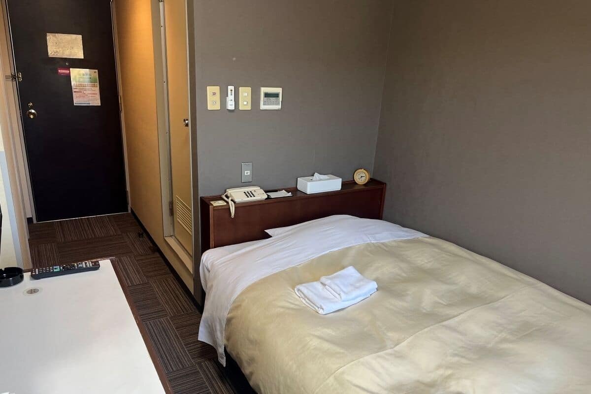 Une chambre d’hôtel avec un lit, une table de chevet avec un téléphone et des mouchoirs, une horloge et une porte avec une pancarte.