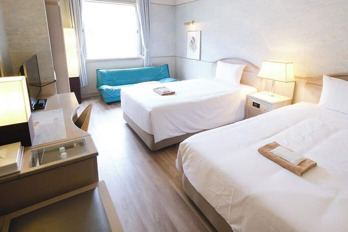 Habitación de hotel con dos camas, un escritorio con lámpara, una silla y una ventana con cortinas.
