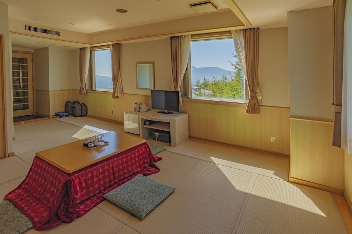 Una habitación con suelo de tatami, una mesa baja con cojines, una televisión y vistas a las montañas a través de las ventanas.