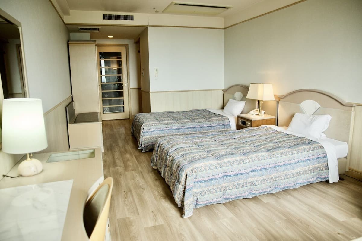 Habitación de hotel con dos camas, un escritorio, una silla, una lámpara y una puerta que da a un pasillo.