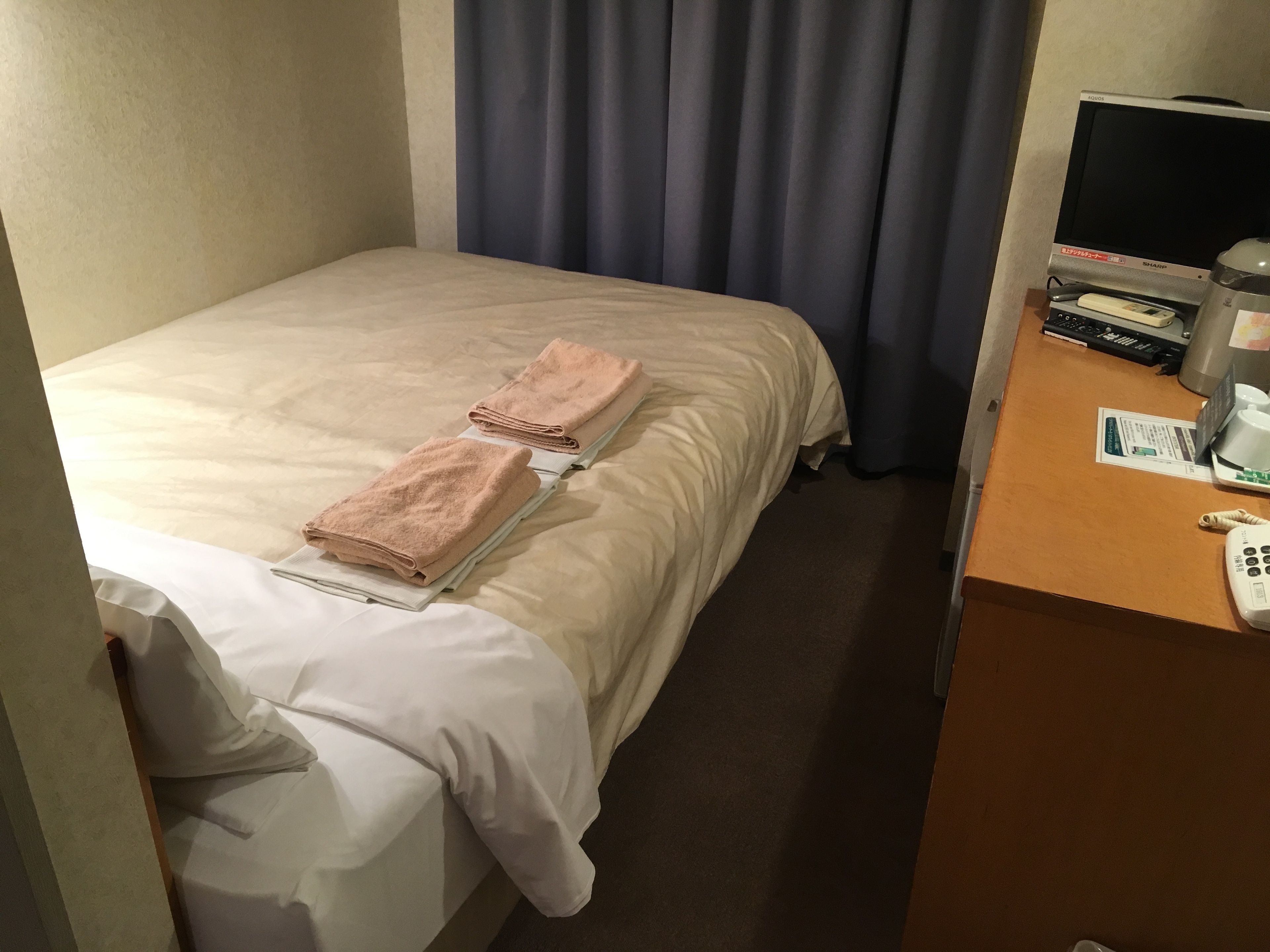 A hotel room with a bed, two towels, a desk with a phone, a monitor, and a coffee maker.