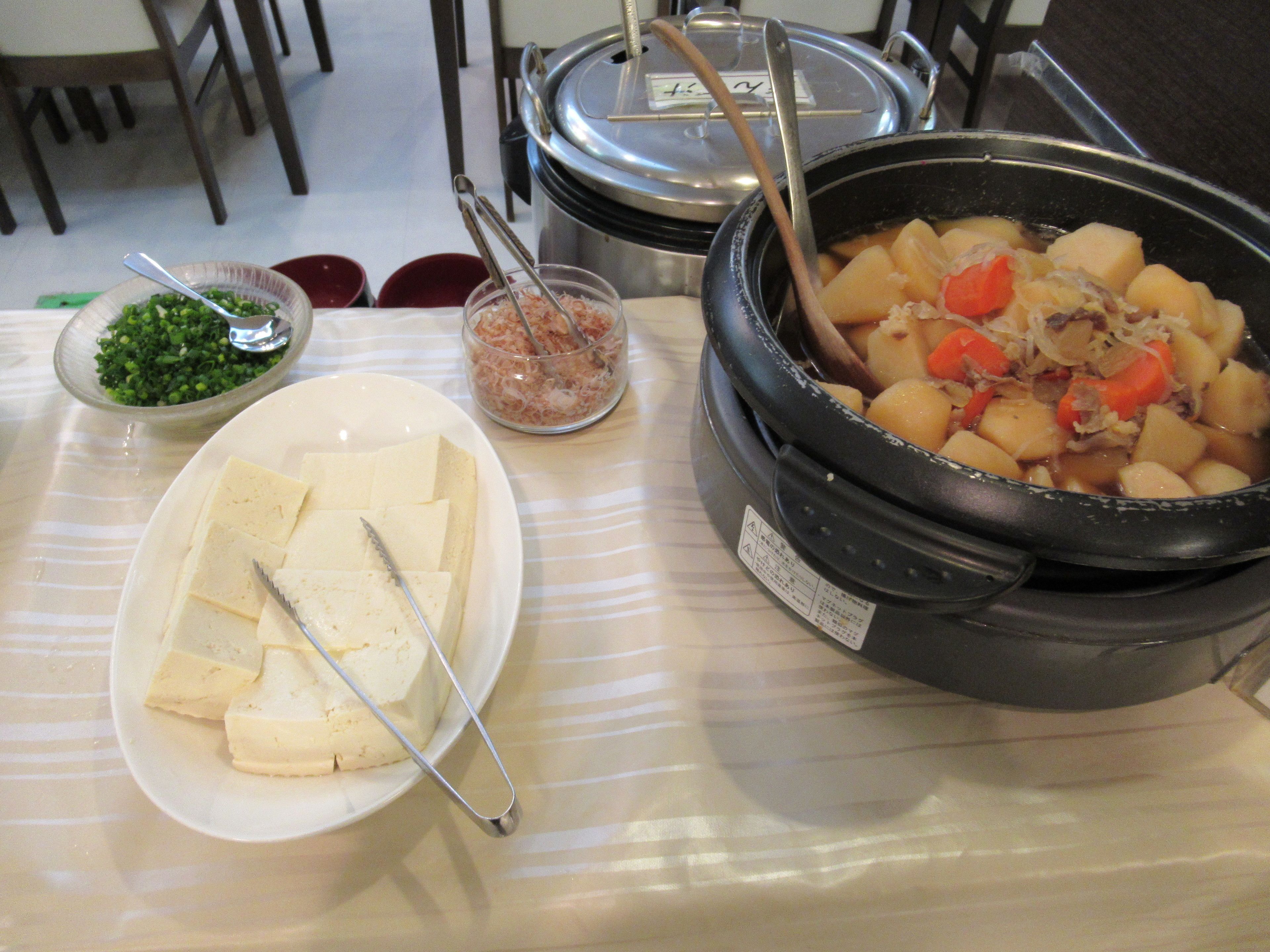 A buffet table with various dishes including a pot of stew, a plate of tofu, and a bowl of green vegetables.