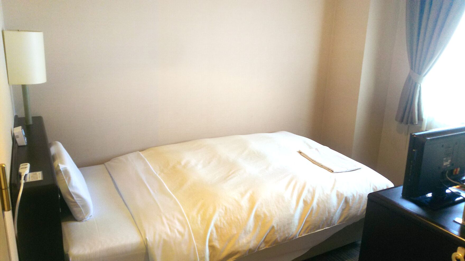 A small, neatly made bed with a white comforter and pillows, a bedside table with a lamp, and a television on a stand.