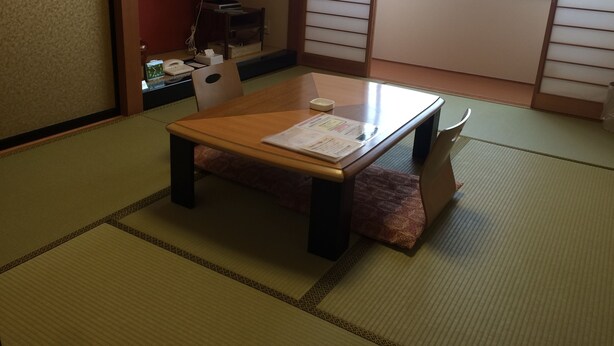 A traditional Japanese room with tatami flooring, a low wooden table with a menu, a chair, and a small desk with a telephone and tissues. - Hotel Crown Hills Takaoka (Takaoka)