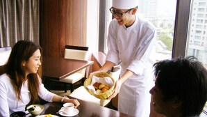 A hotel dining area with a table set for three, a waiter serving food, and a view of the city through large windows. - Resort Capsule Sakuragicho (Yokohama)