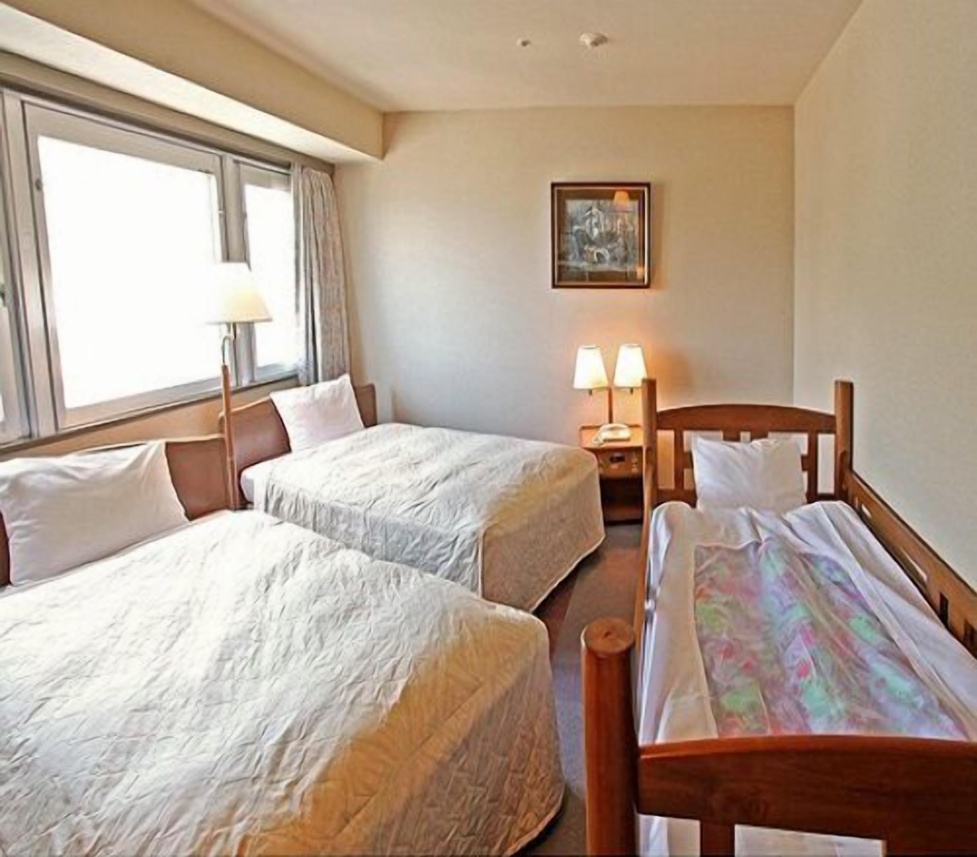 Quarto de hotel com duas camas de solteiro, mesa de cabeceira com luminárias, um quadro na parede e janela com cortinas.