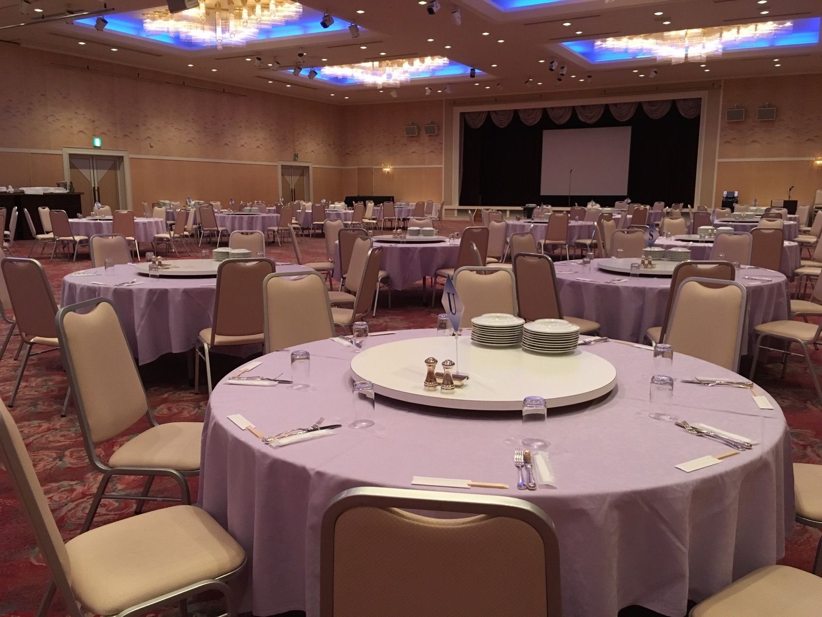 Uma sala de banquetes com mesas redondas cobertas por toalhas roxas, dispostas em fileiras. Cada mesa tem uma pilha de pratos, talheres e um copo. O salão possui um palco com uma tela, e as paredes são decoradas com um padrão texturizado.