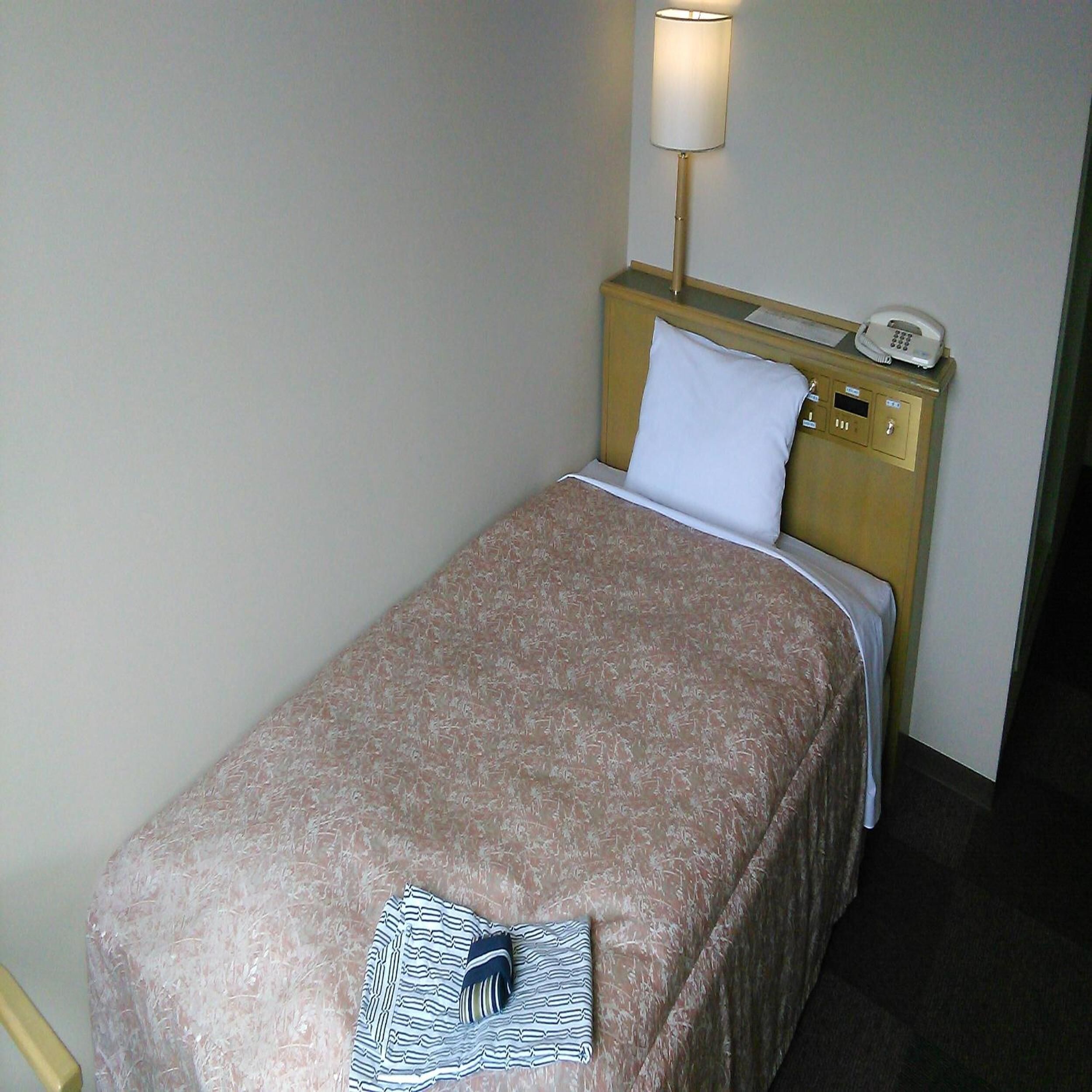 A single bed with a patterned bedspread, a pillow, a bedside lamp, and a telephone on the nightstand.