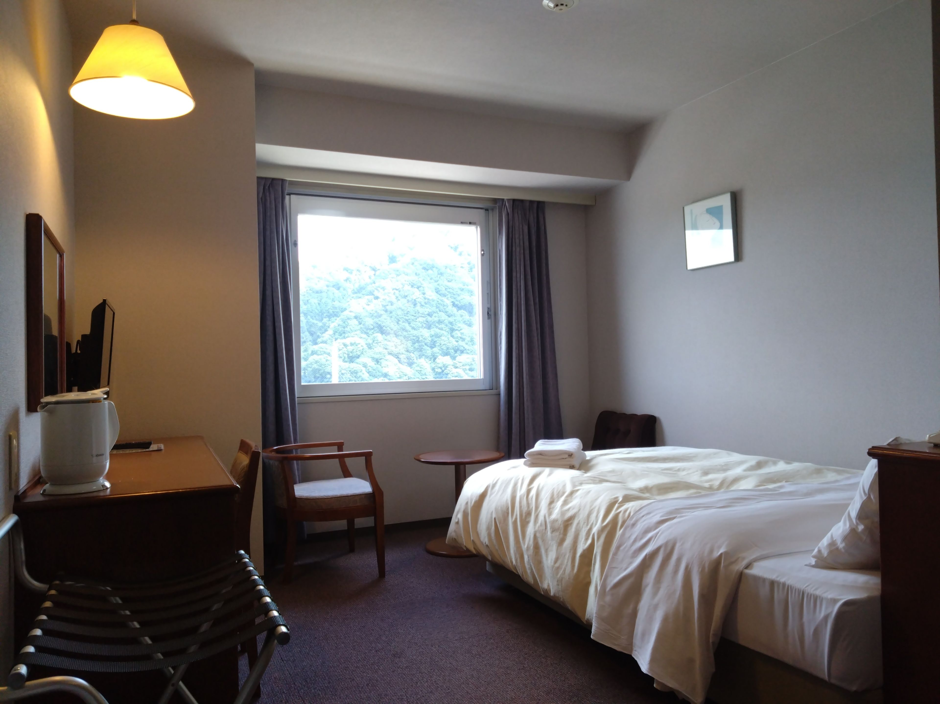 Quarto de hotel com cama, cadeira, escrivaninha, luminária e janela com cortinas.