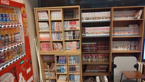 Una librería con numerosas estanterías llenas de manga y libros, una máquina expendedora y una pequeña mesa.