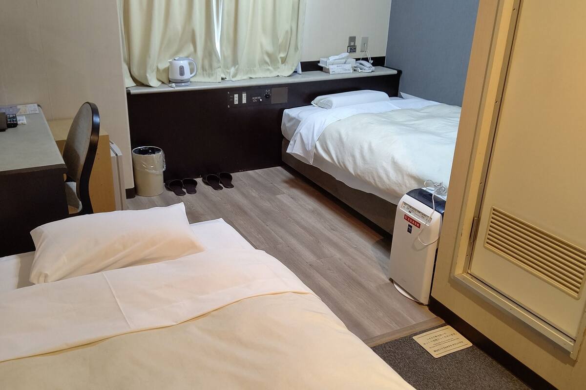 Une chambre d’hôtel comprenant un lit, un bureau avec un ordinateur, une chaise, une poubelle et une table de chevet avec une bouilloire et un téléphone.