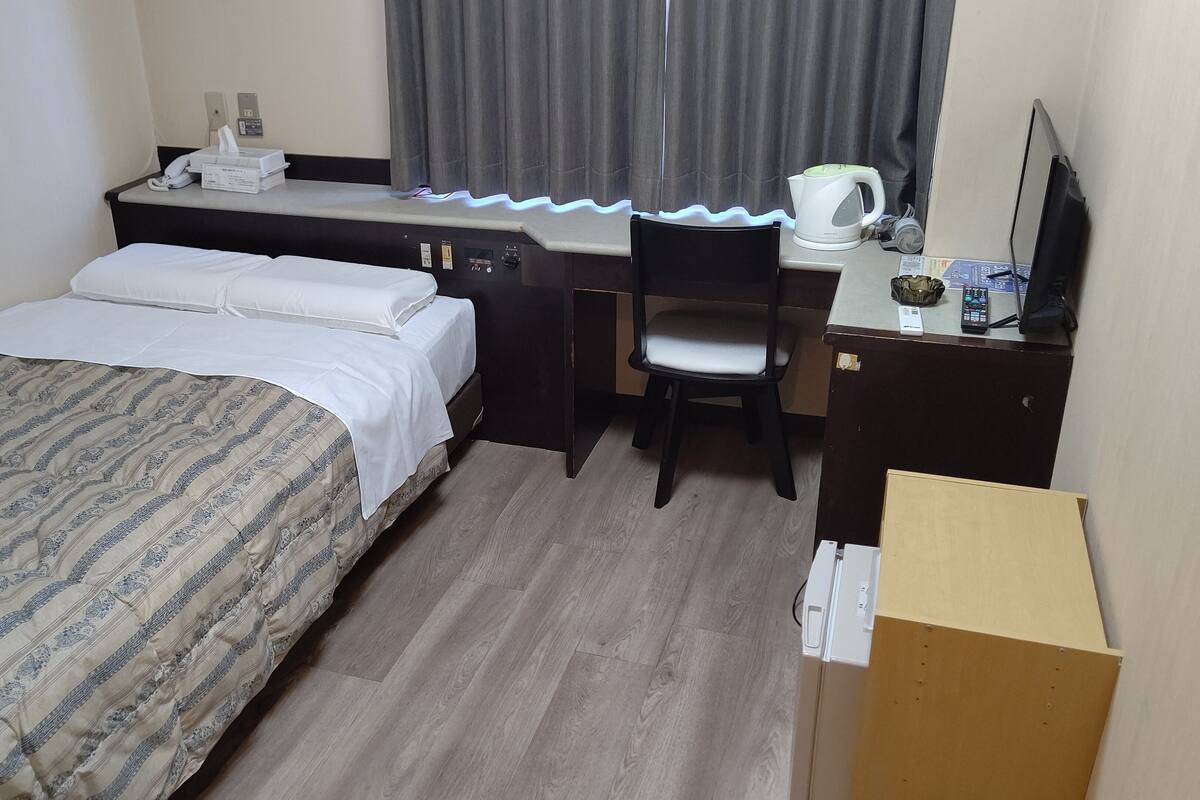 Une chambre d’hôtel équipée d’un lit, d’un bureau, d’une chaise, d’une bouilloire et d’un petit placard.