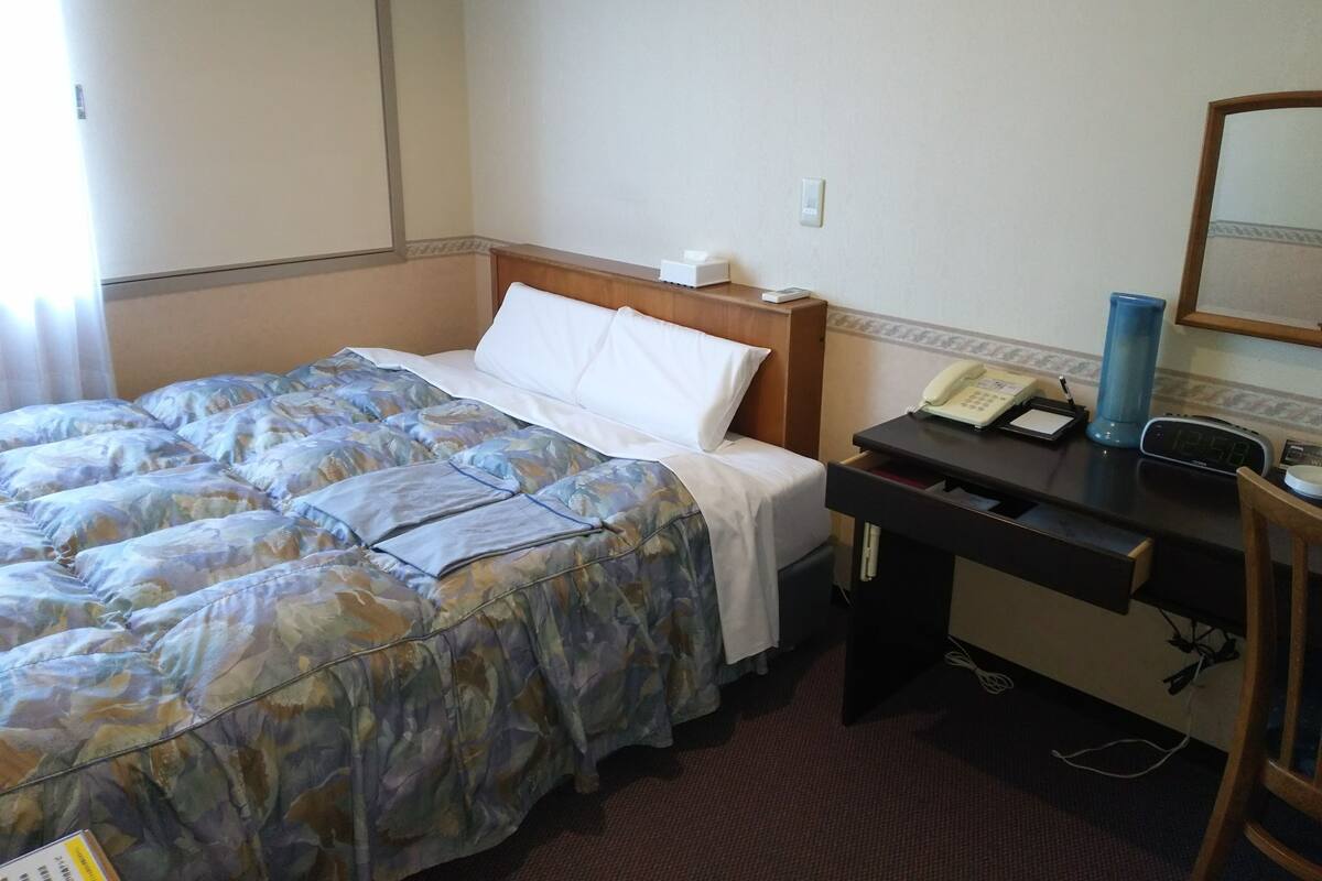 A hotel room with a bed, a desk with a telephone and a clock, and a chair.
