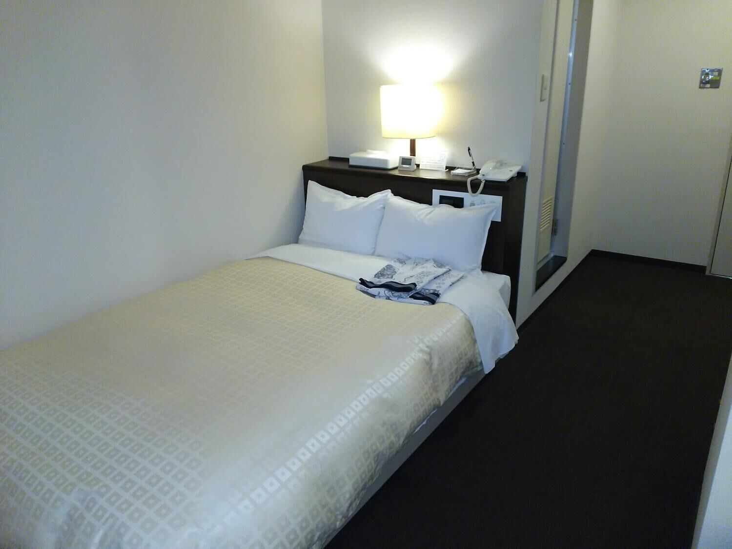 Habitación de hotel con una cama individual, una mesita de noche con lámpara, teléfono y una pequeña mesa con bloc de notas y bolígrafo.
