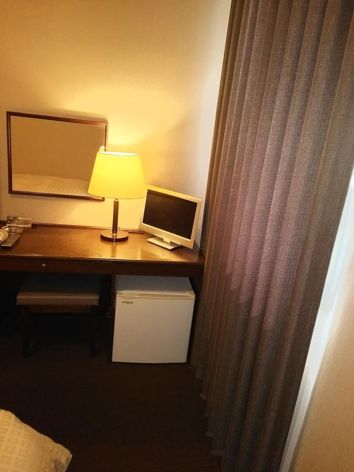 A hotel room with a desk, a lamp, a small refrigerator, a bed, and a window with curtains.