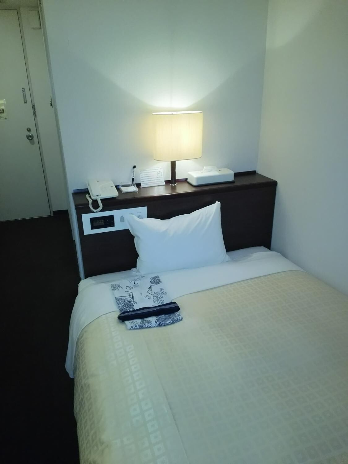A hotel room with a bed, a bedside table with a lamp, a phone, and a sign.