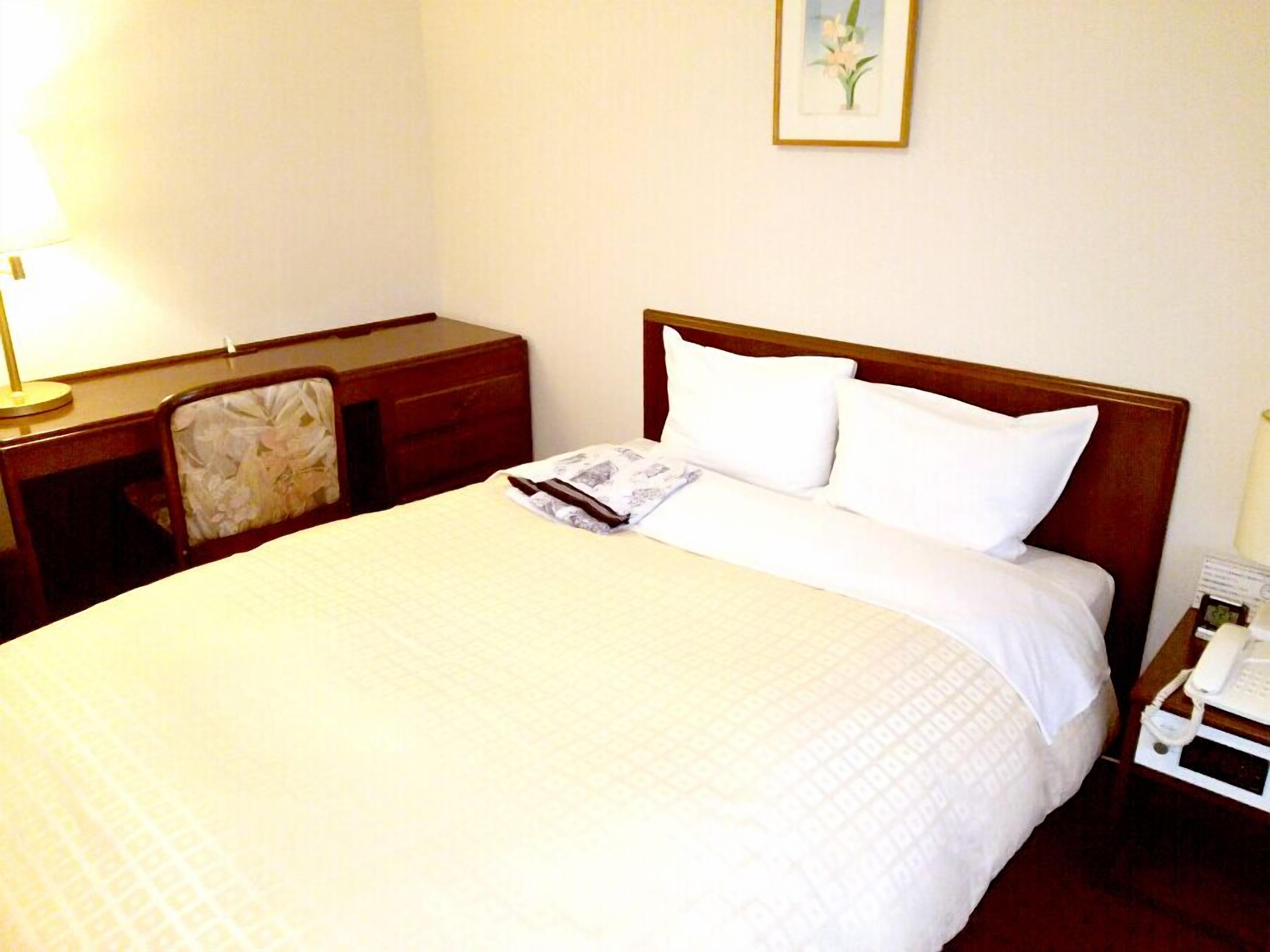 Una cama bien hecha con sábanas blancas, una mesita de noche con lámpara, una silla y un cuadro en la pared.