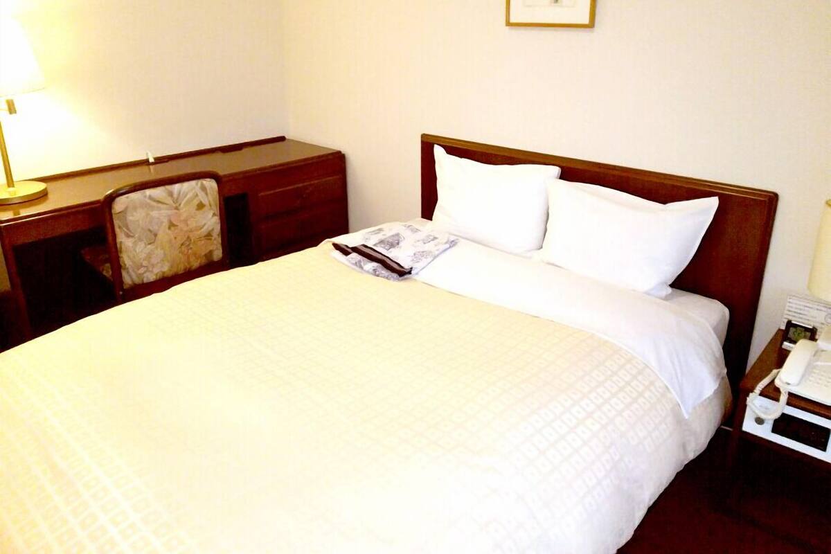 Una cama bien hecha con sábanas blancas, una mesita de noche con lámpara, una silla y un cuadro en la pared.