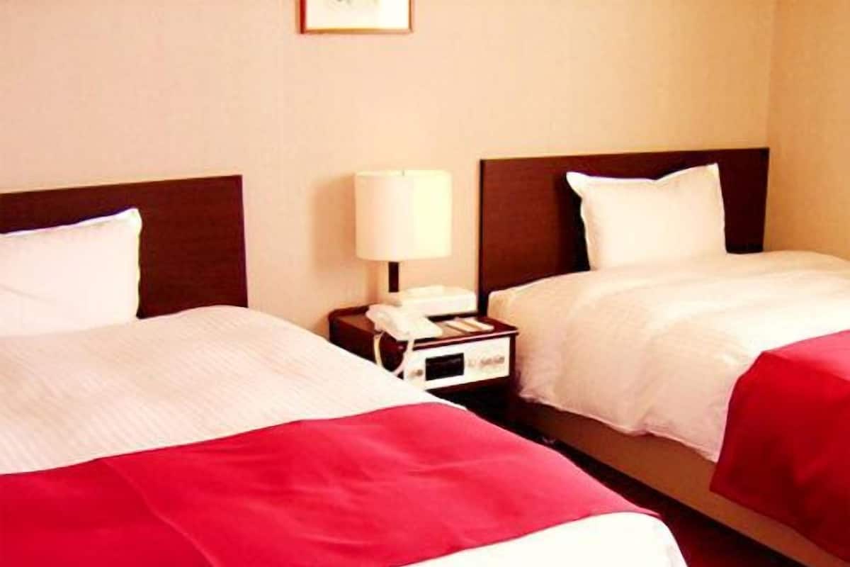 Ein Hotelzimmer mit zwei Betten, jeweils mit weißer Bettwäsche und roten Akzenten. Ein Nachttisch mit Lampe und Telefon ist vorhanden.