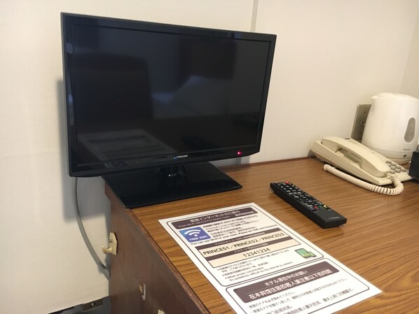 A hotel room with a flat-screen TV, a telephone, and a remote control on a wooden desk.