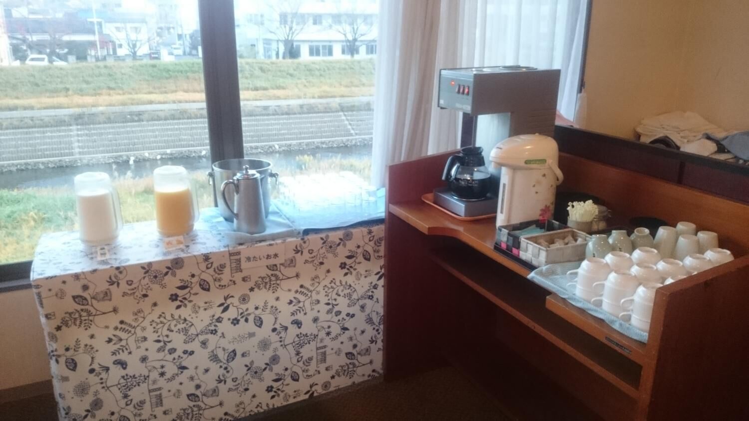 A hotel room with a window view of a railway track and greenery. There is a small table with a coffee maker, a pitcher, and cups.