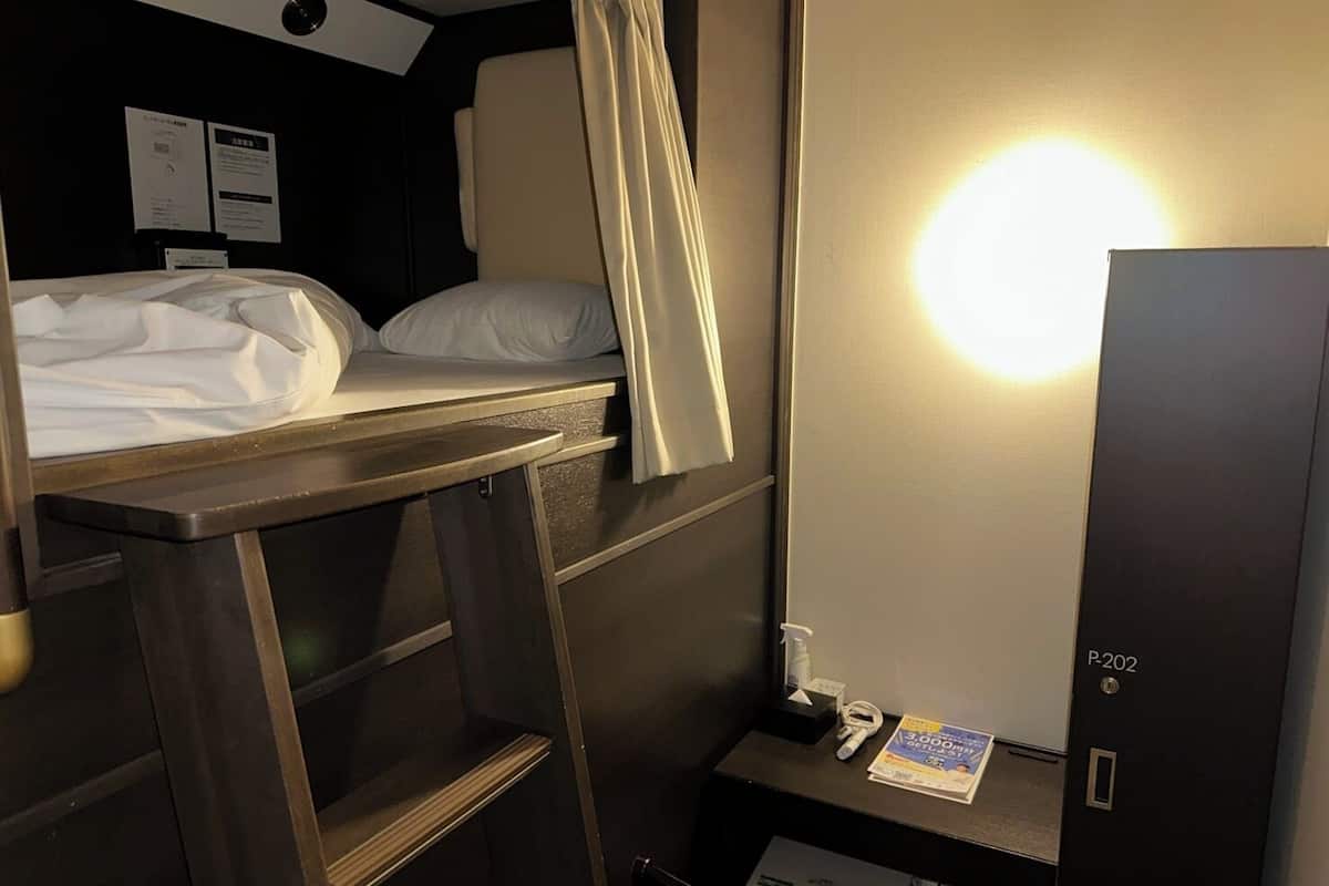 Habitación de hotel pequeña con literas, una mesita de noche con lámpara, un espejo y un escritorio pequeño con silla.