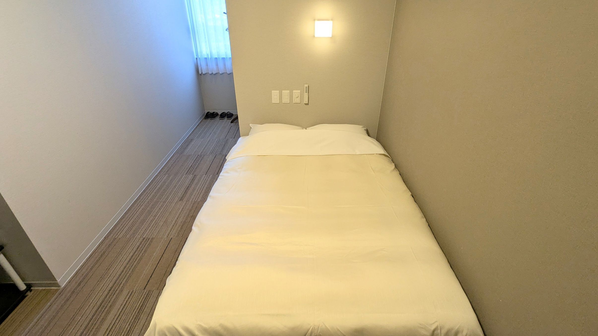 Una cama pequeña y ordenada en una habitación estrecha con ventana, una lámpara de pared y un zapatero.