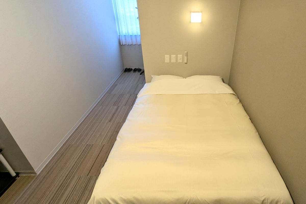 Una cama pequeña y ordenada en una habitación estrecha con ventana, una lámpara de pared y un zapatero.
