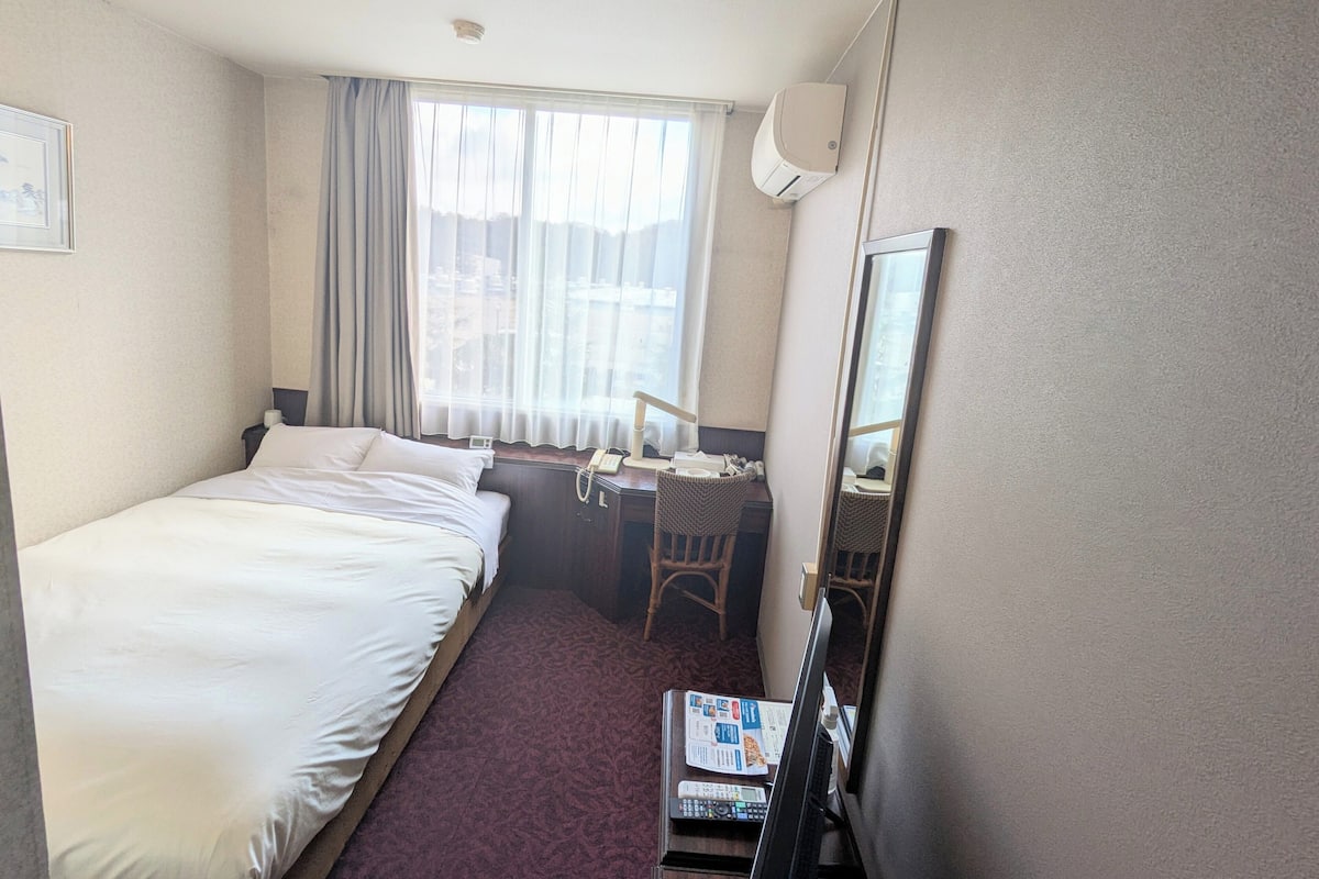 Una habitación de hotel con cama, escritorio con silla, ventana con cortinas, espejo y aire acondicionado de pared.