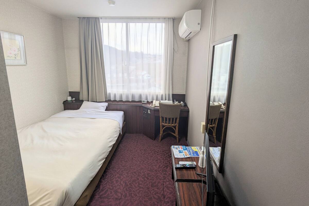 Habitación de hotel con cama, escritorio con silla, espejo, ventana con cortinas y aire acondicionado instalado en la pared.