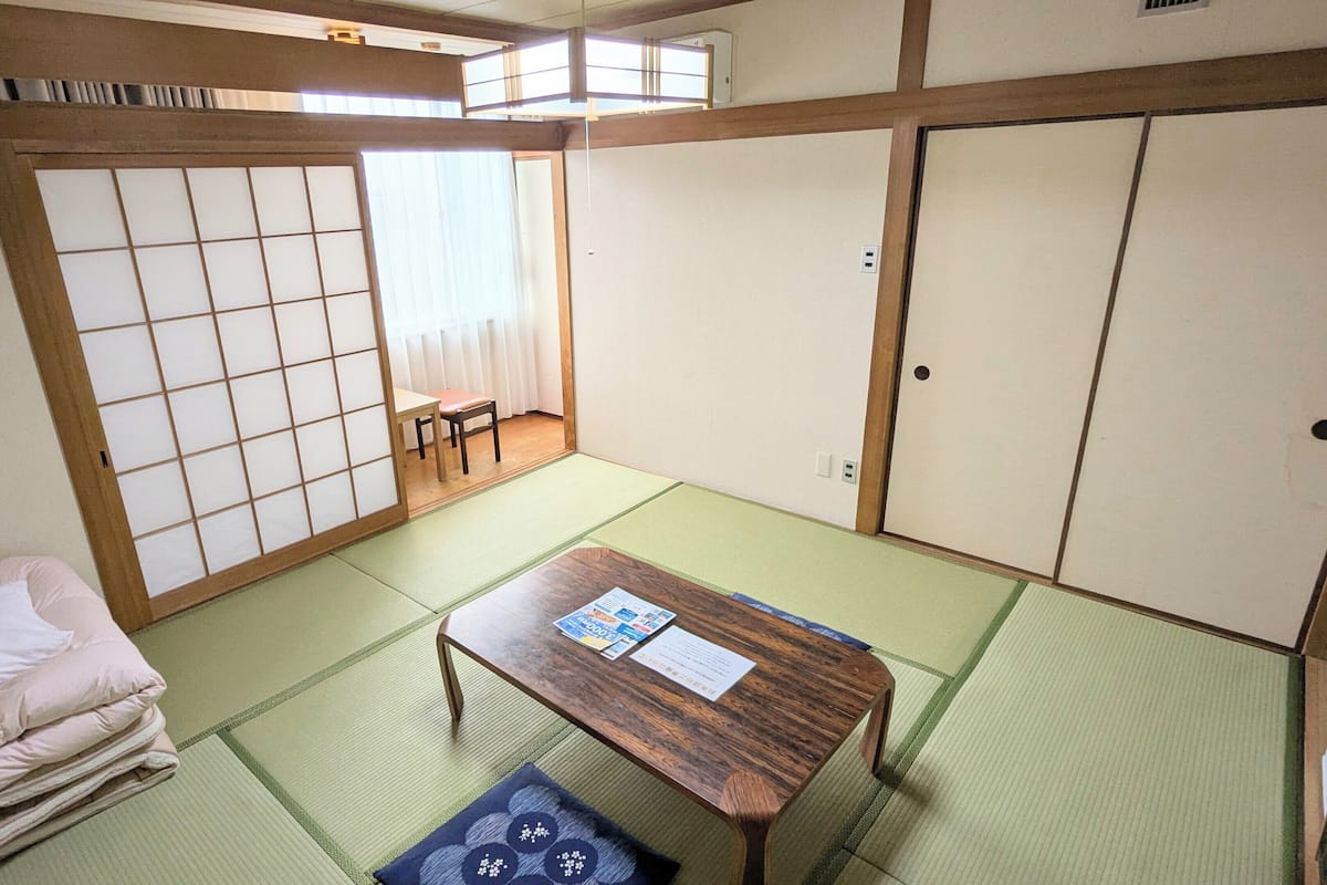 Una habitación tradicional japonesa con suelo de tatami, una mesa de madera baja, un futón como cama, puertas correderas y una ventana con cortinas.